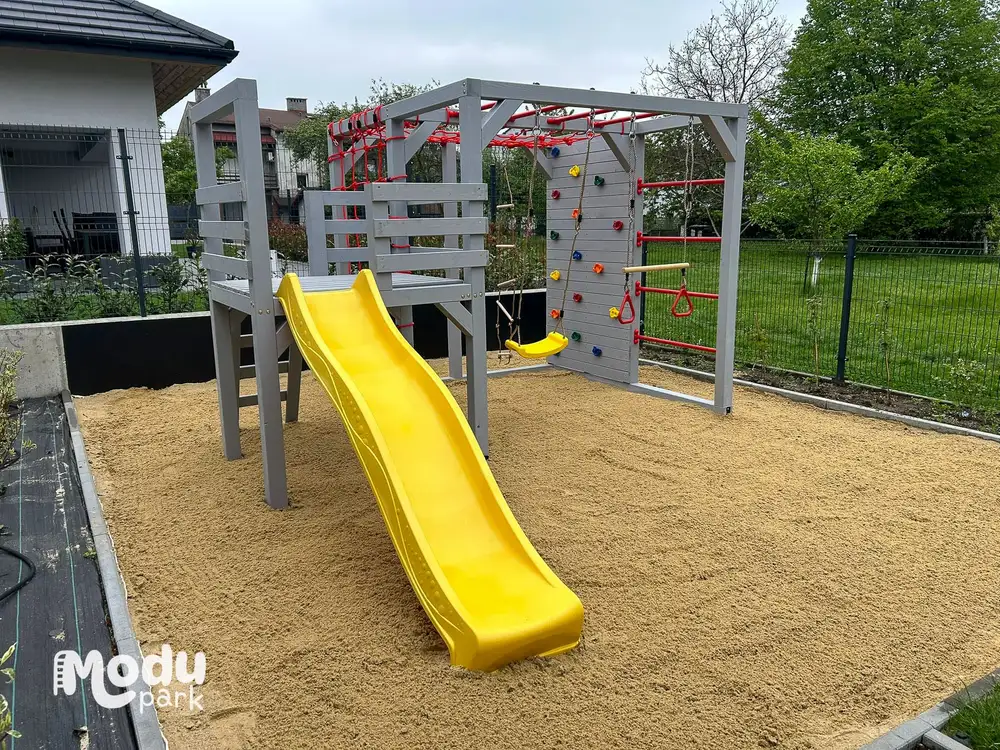 Dziecko zjeżdża ślizgiem z drewnianego placu zabaw ModuPark w ogrodzie, obok huśtawka i podest